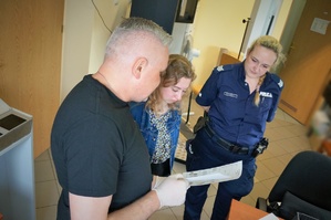 Nieumundurowany policjant - technik kryminalistyki trzyma kartę daktyloskopijną, nad którą stoi praktykantka i policjantka w mundurze