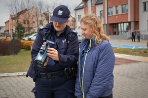 Policjantka z praktykantką stoją na tle komendy. Policjantka trzyma urządzenie do pomiaru prędkości.