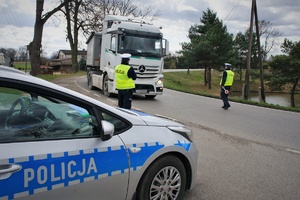 Na pierwszym planie radiowóz, na drugim planie, na jezdni policjanci ruchu drogowego zatrzymują do kontroli samochód ciężarowy