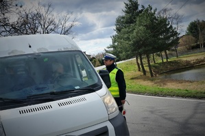 Policjant ruchu drogowego stoi przy kierowcy samochodu w białym kolorze. W tle widać jezdnię i drzewa