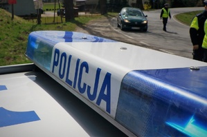 Na pierwszym planie listwa sygnalizacyjna radiowozu. Na drugim planie widać kontrolujących policjantów i zatrzymany do kontroli samochód osobowy