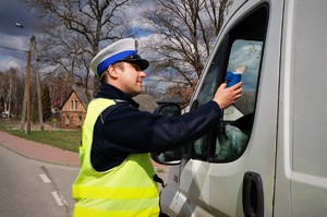 Policjant ruchu drogowego stoi przy kierowcy zatrzymanego do kontroli samochodu. W wyciągniętej w kierunku kierowcy dłoni trzyma urządzenie do pomiaru trzeźwości