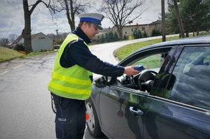 Policjant ruchu drogowego stoi przy kierowcy zatrzymanego do kontroli samochodu. W wyciągniętej w kierunku kierowcy dłoni trzyma urządzenie do pomiaru trzeźwości