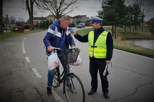 Na poboczu jezdni  policjant ruchu drogowego stoi przy mężczyźnie na rowerze trzymając przy jego ustach urządzenie do pomiaru trzeźwości