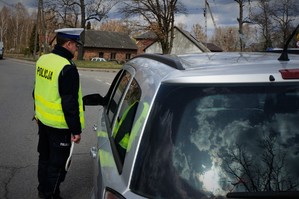 Policjant ruchu drogowego stoi przy kierowcy zatrzymanego do kontroli samochodu. W wyciągniętej w kierunku kierowcy dłoni trzyma urządzenie do pomiaru trzeźwości