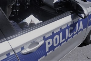 Fragment radiowozu, w którym siedzi policjant dokumentujący czynności służbowe. Na drzwiach pojazdu napis: POLICJA