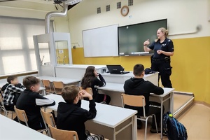 Policjantka na prelekcji w sali lekcyjnej, stoi pod tablicą przodem do uczniów, którzy siedzą w ławkach. W tle ściana w kolorze żółtym