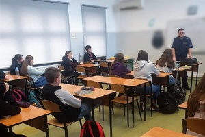 Policjant na prelekcji w sali lekcyjnej, stoi pod tablicą przodem do uczniów, którzy siedzą w ławkach