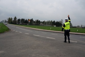 Na jezdni stoi policjant ruchu drogowego z prawą ręką uniesioną do góry, zatrzymuje do kontroli samochód.