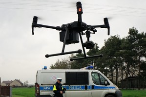 W górnej części zdjęcia uniesiony nad ziemią dron. Na drugim planie, poniżej policyjny radiowóz przy którym stoi policjant ruchu drogowego