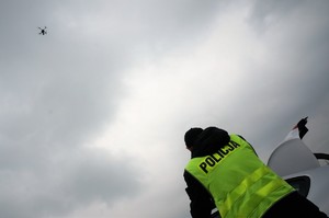 Na tle zachmurzonego nieba, w prawym dolnym rogu widać fragment policjanta w odblaskowej kamizelce z napisem POLICJA, w lewym górnym rogu w oddali widać drona