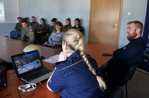 Na pierwszym planie dwoje policjantów siedzi za stołem, przed policjantką leży laptop. Uczniowie w mundurach siedzą na krzesłach ustawionych w rzędy pod ścianą