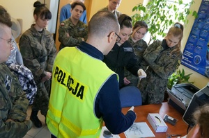Zajęcia u technika kryminalistyki. Policjant w odblaskowej kamizelce prezentuje uczniom techniki pobierania śladów. Uczniowie w mundurach stoją wokół policjnata