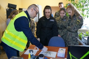 Zajęcia u technika kryminalistyki. Policjant w odblaskowej kamizelce prezentuje uczniom techniki pobierania śladów. Uczniowie w mundurach stoją wokół policjnata