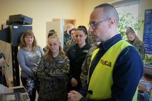 Na pierwszym planie technik kryminalistyki w odblaskowej kamizelce z napisem POLICJA. Na drugim planie uczniowie w mundurach i wychowawczyni