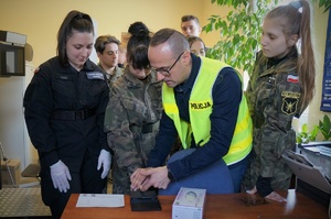 Zajęcia u technika kryminalistyki. Policjant w odblaskowej kamizelce prezentuje uczniom techniki pobierania śladów. Uczniowie w mundurach stoją wokół policjanta