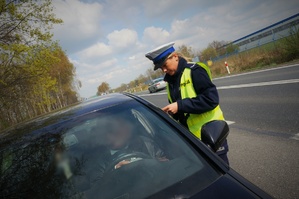 Policjantka ruchu drogowego na poboczu jezdni stoi przy kierowcy zatrzymanego do kontroli pojazdu. W tle widać jezdnię, drzewa i niebo