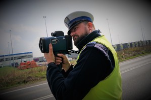 Policjant ruchu drogowego na tle jezdni stoi trzymając laserowe urządzenie do pomiaru prędkości