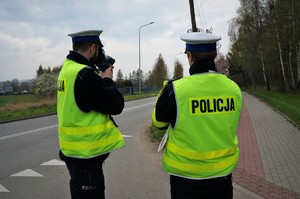 Policjanci ruchu drogowego stoją na poboczu jezdni, jeden z nich mierzy prędkość pojazdów. Na odblaskowej kamizelce jednego policjanta widać napis POLICJA. W tle jezdnia, niebo i drzewa