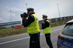 Policjanci ruchu drogowego stoją przy radiowozie na poboczu jezdni. Policjant na pierwszym planie trzyma laserowe urządzenie do pomiaru prędkości