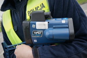 Zbliżenie na laserowe urządzenie do pomiaru prędkości, które trzyma policjant. Na odblaskowej kamizelce widać napis POLICJA