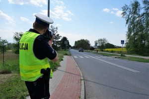 Na pierwszym planie policjant ruchu drogowego stoi na chodniku przy jezdni z laserowym miernikiem prędkości. Na drogim planie widać przejście dla pieszych.