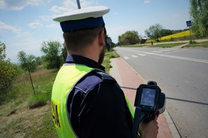 Na pierwszym planie policjant ruchu drogowego trzyma w ręku laserowy miernik prędkości. Na drugim planie widać jezdnię i przejście dla pieszych