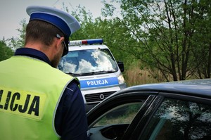 Policjant ruchu drogowego podczas kontroli drogowej stoi przy ciemnym samochodzie, którego fragment widać w prawym dolnym rogu. Na drugim planie stoi radiowóz i widać też drzewa