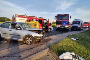 Na pierwszym planie na jezdni stoi rozbity samochód. Na drugim planie pojazdy służb ratunkowych