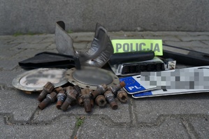 Zabezpieczone przez Policję części samochodowe i tablice rejestracyjne leżą na ziemi. Na częściach leży opaska z napisem POLICJA