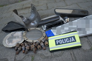 Zabezpieczone przez Policję części samochodowe i tablice rejestracyjne leżą na ziemi. Na częściach leży opaska z napisem POLICJA