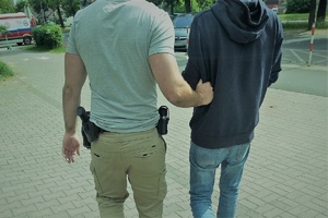 Nieumundurowany policjant kryminalny trzyma pod rękę zatrzymanego mężczyznę. W tle widać chodnik. Na drugim planie stoi karetka pogotowia