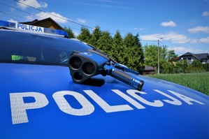 Laserowy miernik prędkości leży na masce radiowozu. Na pierwszym planie widać napis POLICJA. W tle widać zielone krzewy i niebo