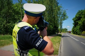 Policjant ruchu drogowego stoi przy jezdni z laserowym miernikiem prędkości. W tle widać jezdnię przy której rosną drzewa.