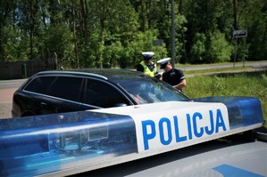 Na pierwszym planie listwa sygnalizacyjna radiowozu, na której widać napis POLICJA. Na drugim planie policjanci stoją przy zatrzymanym do kontroli drogowej ciemnym samochodzie osobowym. W tle widać jezdnię i drzewa