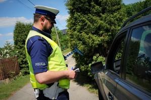 Policjant ruchu drogowego stoi przy zatrzymanym do kontroli samochodzie trzymając w ręku dokumenty od kierowcy. W tle widać szutrową drogę i drzewa