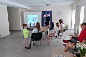 Duża sala Centrum Usług Społecznych w Lędzinach. Na pierwszym planie widać siedzące na krzesłach dzieci. Na drugim planie przy monitorze z prezentacją stoi policjant prowadzący prelekcję