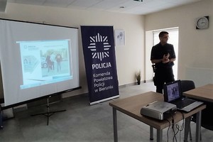 Sala Centrum Usług Społecznych w Lędzinach. Na zdjęciu widać policjanta prowadzącego prelekcję który stoi przy banerze Komendy Powiatowej Policji w Bieruniu z logo Policji i ekranie z multimedialną prezentacją