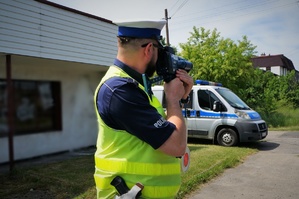 Policjant ruchu drogowego stoi z miernikiem prędkości przyłożonym do twarzy. W tle stoi zaparkowany radiowóz