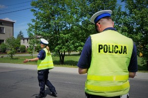 Policjanci ruchu drogowego: po lewej stronie na jezdni policjantka zatrzymuje do kontroli samochód, po prawej stoi policjant w odblaskowej kamizelce, na plecach widoczny napis POLICJA