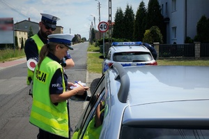 Policjanci ruchu drogowego stoją przy zatrzymanym do kontroli samochodzie w kolorze szarym, przy oknie kierowcy. W tle widać zaparkowany przy poboczu jezdni radiowóz