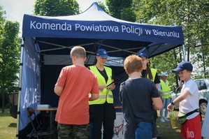 Na pierwszym planie stoją trzej chłopcy odwróceni przodem do uczennicy z Młodzieżowej Służby Ruchu Drogowego w odblaskowej kamizelce. Na drugim planie widać policyjny namiot profilaktyczny