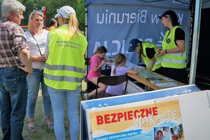 Na pierwszym planie widać fragment banera z napisem: Bezpieczne wakacje. W tle profilaktyczny namiot Policji a w nim dwie uczennice i opiekunka Młodzieżowej Służby Ruchu Drogowego. W namiocie przy stoliku siedzą dzieci