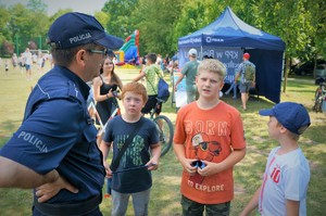 Na pierwszym planie stoi policjant a przed nim trzech chłopców. W tle widać policyjny namiot profilaktyczny i boisko na Mini Arboretum
