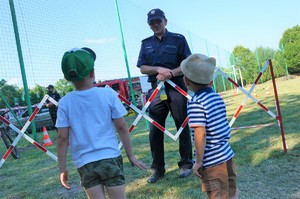 Na pierwszym planie dwaj mali chłopcy, którzy stoją przodem do policjanta. W tle widać trawę i niebo