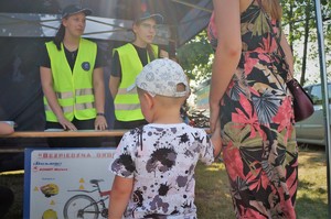 Na pierwszym planie przy policyjnym stoisku profilaktycznym stoi mały chłopiec, którego trzyma za rękę opiekunka w kolorowej sukience. Na drugim planie pod namiotem, za stolikiem stoją dwie uczennice w odblaskowych kamizelkach