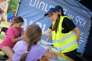 W centralnej części zdjęcia uczennica z Młodzieżowej Służby Ruchu Drogowego w odblaskowej kamizelce stoi pod namiotem profilaktycznym Policji, spogląda na dziewczynki siedzące za stolikiem