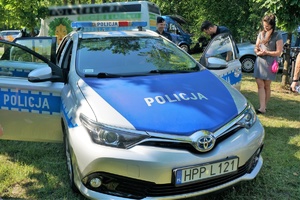 Na pierwszym planie widać od przodu policyjny radiowóz, w którym są otwarte drzwi. Przy radiowozie stoją uczestnicy festynu