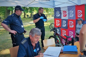 Punkt znakowania rowerów. Na pierwszym planie za stolikiem siedzi strażnik miejski, który wypisuje dokumentację. Na drugim planie stoi Komendant Powiatowy Policji w Bieruniu a obok niego strażnik miejski, który znakuje rowery. Na tle kolorowej ścianki namiotu stoi rower a przy nim chłopiec
