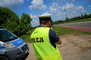 Policjant ruchu drogowego stoi przodem do jezdni dokonując pomiaru prędkości pojazdów. Po lewej stronie widać fragment radiowozu